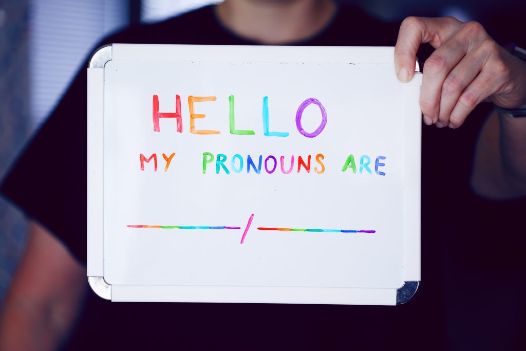 Eine Person hält eine kleine weiße Tafel, auf der in Regenbogenfarben Folgendes steht: Hello my pronouns are. Datunter sind leere Striche, getrennt durch einen Schrägstrich. Sie dienen als Platzhalter, um Pronomen einzutragen.