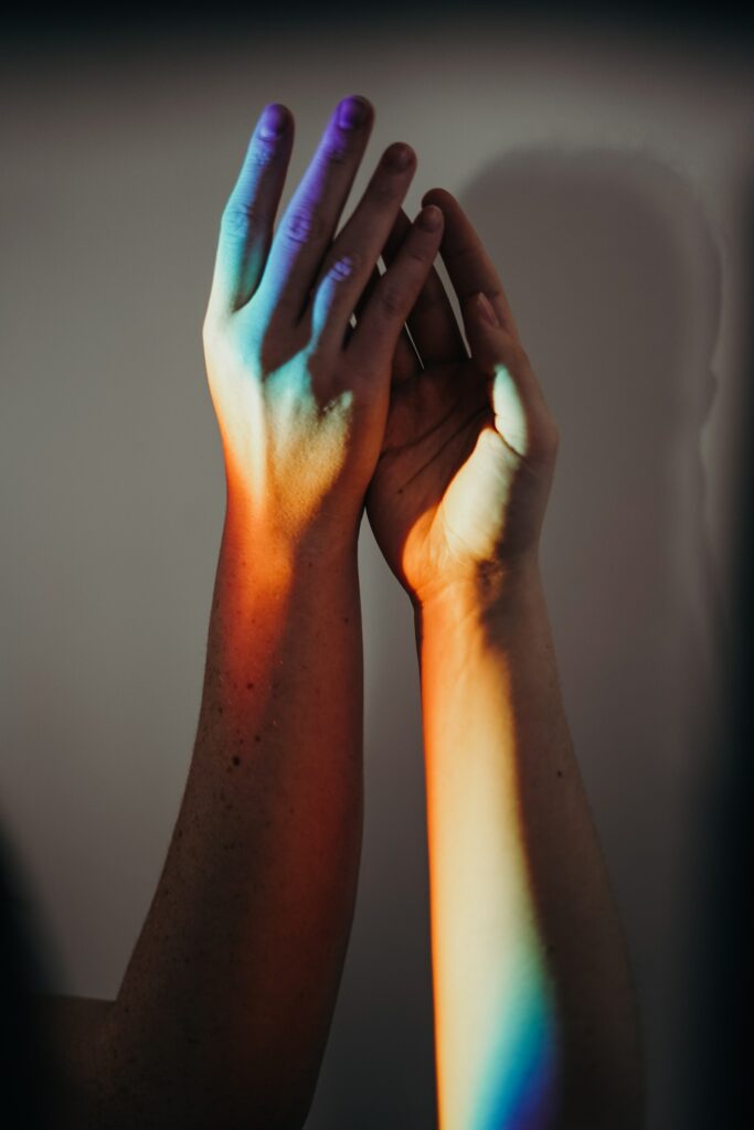 Foto von zwei Händen die sich berühren. Angestrahlt von einem Regenbogen.